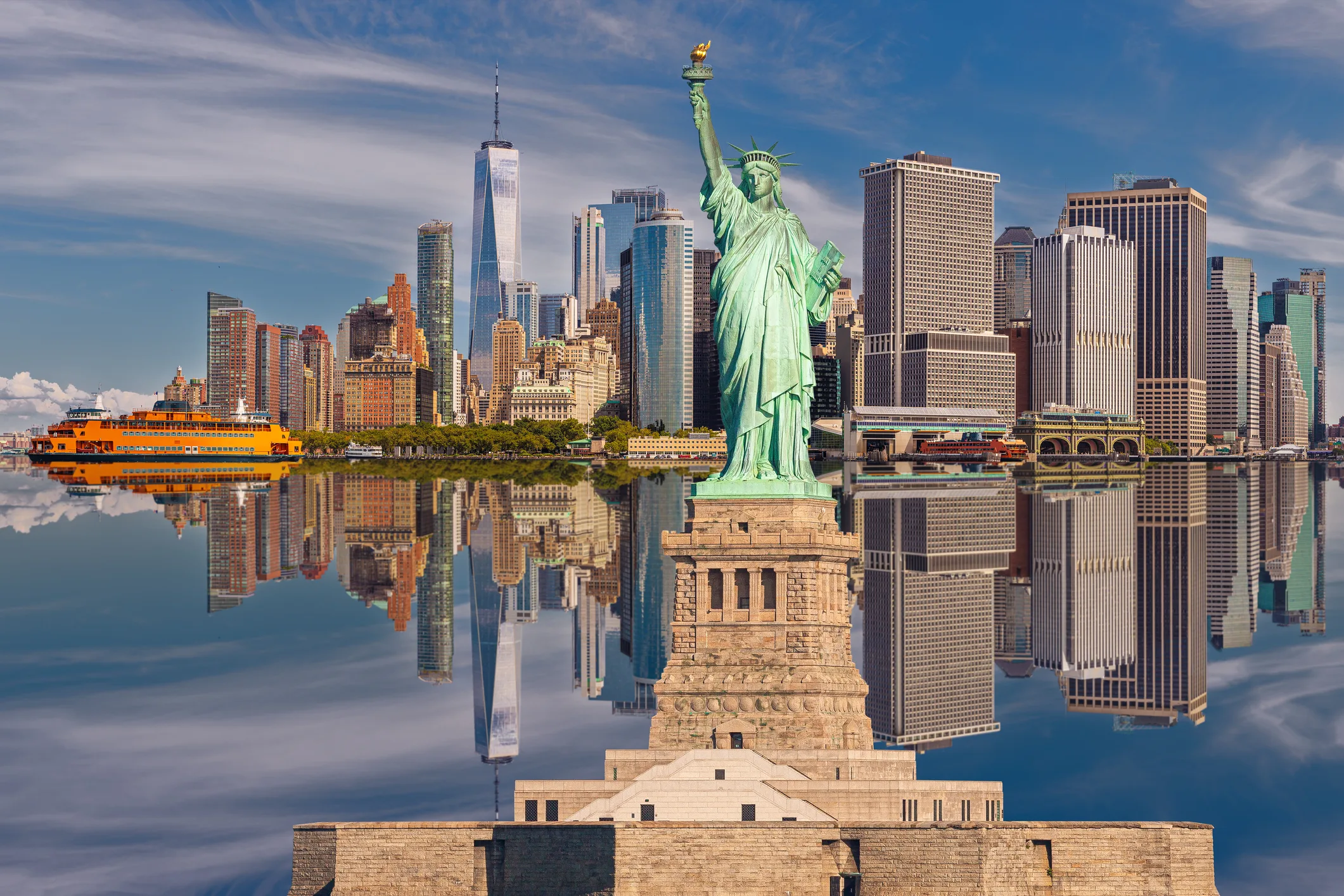 Statue of Liberty and New York City Skyline with Manhattan Financial District, Battery Park,  World Trade Center, Staten Island Ferry and Blue Sky with Clouds All Reflected in Water of New York Harbor. High Resolution Stitched Panoramic image with 2:1 image aspect ratio. Canon EOS 6D Full Frame Sensor Camera and Canon EF 85mm f/1.8 USM Prime Lens.