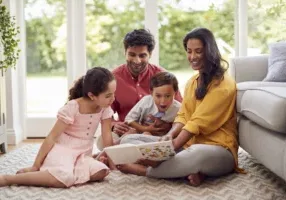 Family At Home Sitting On Floor In Lounge Reading Book Together